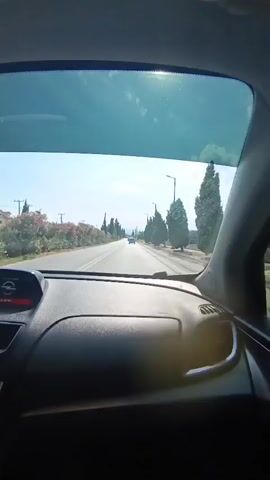 The video shows a view from the inside of a car looking out the windshield. The car is driving on a paved road with trees and bushes lining both sides. There are streetlights visible along the road. The sun is shining brightly, creating lens flare on the windshield. There is a dark dashboard and steering wheel in the foreground. A red light is visible on the dashboard. The road stretches into the distance, with another vehicle visible ahead. The sky is blue with some clouds. The overall scene suggests a sunny day drive in a rural or suburban area.