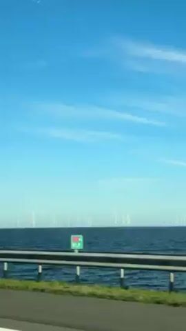 The video shows a view from a moving vehicle, looking out over a body of water with a wind farm. The sky is blue with thin, wispy clouds. A metal guardrail runs along the edge of the road beside the water. Many wind turbines are visible in the distance, standing in the water. The road is asphalt, and a strip of green grass is visible between the road and the guardrail. The camera is stable.