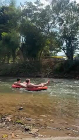 Two people float in red inflatable tubes in a river, surrounded by lush green vegetation. The riverbank, visible in the foreground, consists of sand and small rocks. The water is clear, with visible currents. In the background, a simple structure partially covered with fabric or netting is visible through the trees. The overall setting is a natural, tropical environment.