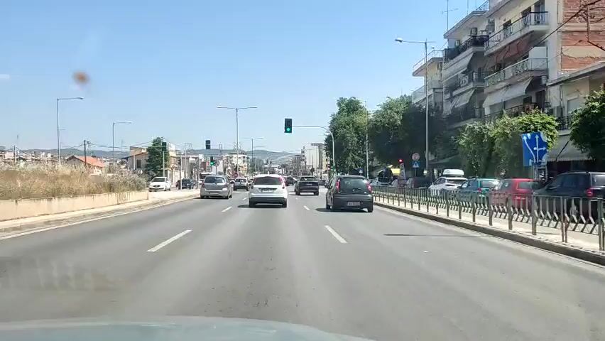 A dashcam perspective captures a vehicle traveling down a multi-lane road on a clear, sunny day. The road is asphalt with white lane markings. Multiple cars are visible in both directions. On the left side of the road, there's a dirt area with some sparse vegetation and residential buildings in the distance. On the right, there are buildings, some with stores on the ground floor, and a sidewalk with a metal barrier. Traffic lights are visible at intersections, and some trees line the right side of the road. A few brown spots are visible on the windshield, possibly smudges or glare.