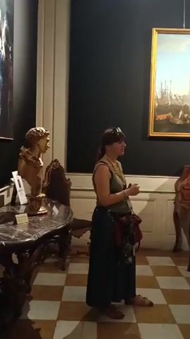 In this video, a woman is standing in a museum, possibly giving a tour. She is wearing a green sleeveless top, a long dark skirt, and sandals. A dark red bag with a green scarf is slung across her body. She is gesturing with her hands as she speaks. Behind her, a large painting hangs on the wall depicting a busy harbor scene with sailing ships and people. In front of the painting is an ornate wooden desk with many small drawers. To the left of the woman, a bust on a pedestal is visible, along with a dark marble-topped table and a wooden chair. The floor is tiled in a checkered pattern of light and dark squares. The lighting suggests it is daytime, and the atmosphere appears to be educational and engaging. The overall scene is characteristic of a museum or gallery setting.