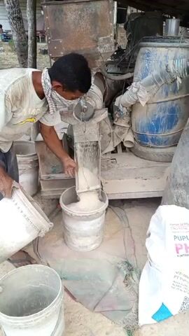 A man wearing a white t-shirt and a checkered cloth mask over his face is operating a rice milling machine. He is standing on the left side of the frame, facing the machine. He is holding a white bucket in his left hand, pouring its contents into the top of the machine, and with his right hand, he is guiding a stream of rice falling from the machine into another white bucket placed below. The machine appears to be a small, portable unit with a blue barrel to its right. The ground is sandy and covered with a patterned fabric underneath the buckets. Several other buckets are visible around the work area. A white bag with text and a blue border is leaning against the machine on the right side of the frame. The background shows trees and vegetation. The video captures the process of rice milling.