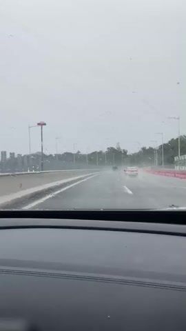 A view from inside a car driving on a highway on a rainy day. Water droplets are visible on the windshield. The road is wet, and spray is coming up from the tires of the car and other vehicles. Several cars are on the road, some appearing to be white or light-colored. There are guardrails and streetlights along the side of the road. Trees and vegetation are visible beyond the guardrails. The sky is overcast.