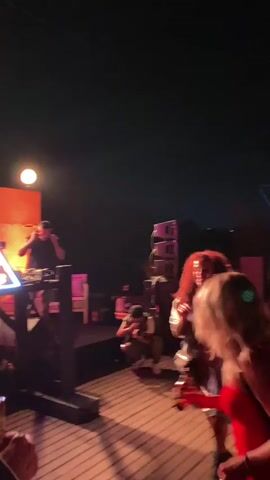 The video shows an outdoor party scene at night. In the foreground, two women are dancing. The woman on the right is wearing a red dress and has blonde hair. The woman on the left has curly red hair and is wearing a shiny, patterned dress. She is holding a drink. In the background, a male DJ is visible behind a table with audio equipment. He is wearing a black t-shirt and a baseball cap. He appears to be playing music. There are stage lights and speakers set up. The overall atmosphere suggests a lively and energetic event.