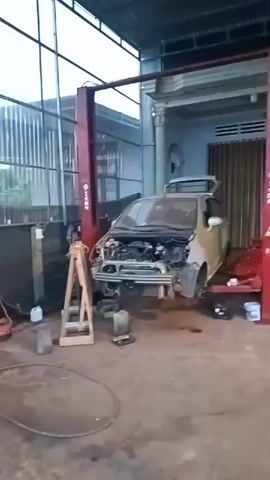 The video shows the inside of an auto repair shop. A two-post lift is centrally positioned in the frame, with a disassembled car raised on it. Several other vehicles, including a teal hatchback and a burgundy van, are parked around the lift. A red motorcycle is parked close to the lift. The shop is open-air, with a corrugated metal roof, and looks to be attached to a house with a white portico. Tools and car parts are scattered on the floor.