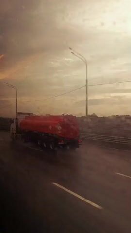 A tanker truck, painted red with hazard symbols and text, drives down a wet highway under a cloudy sky. The truck has multiple axles and a long, cylindrical tank. A white station wagon passes the truck, then another car is visible in the distance. The scene appears to be filmed from inside another vehicle, with water droplets on the window obscuring the view slightly. Streetlights and a barrier line the side of the highway.