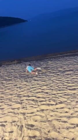 The video shows a person doing sit-ups on a sandy beach. The person is wearing a white shirt and blue shorts. The beach is next to a large body of water, which appears to be a lake or the sea. The sky is dark, indicating it is either dusk or night. The waves on the water are gentle. The sand is light brown and has ripples from the wind. The person is in motion, with their body positioned as if they are in the middle of a sit-up.
