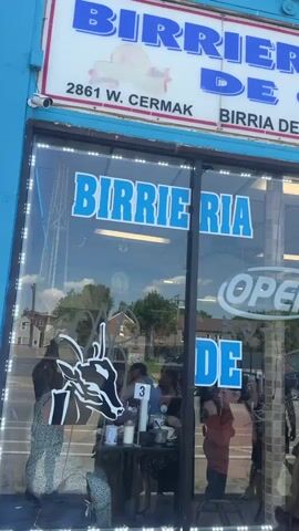 A daytime exterior shot captures a bustling Birrieria La Tapatia De Ocotlan restaurant. The turquoise and white signage is prominently displayed above the storefront, adorned with a depiction of a cow. The restaurant's glass windows feature the name "BIRRIERIA" in large, blue letters. Inside, patrons are seated at tables enjoying their meals. Outside, a diverse group of people is waiting, their interactions and expressions suggesting anticipation. The building itself is brick, and the surrounding environment includes a street scene with houses visible in the background. The overall atmosphere is lively, indicating a popular local eatery.