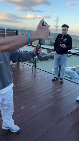 This short video appears to capture a casual gathering on a wooden deck, possibly a rooftop or patio, in Lisi, Georgia. The time of day suggests late afternoon or early evening, with a clear sky and soft lighting. In the foreground, a man wearing a grey hoodie, a black baseball cap, and sunglasses is the primary subject. He is holding a smartphone, seemingly filming or taking a selfie, and also holds a microphone in his left hand. He gestures with his right hand, pointing towards the camera, and appears to be speaking or performing. His attire, including camouflage patterned pants, suggests a relaxed and possibly artistic or musical context.

Behind him, another person wearing a dark jacket, white t-shirt, and a black beanie is visible, standing near a table with some items on it, including clothing items that appear to have logos. Other individuals can be faintly seen in the background, adding to the sense of a social event. A glass of white wine is held up in front of the camera at one point, implying a celebratory or social toast. The overall mood seems energetic and convivial, with the primary subject exuding confidence and engagement. The background reveals buildings and a hilly landscape, consistent with the geographical context of Lisi. Visible text includes a logo on the baseball cap, which appears to be "NY," and text on a blue bracelet worn by the main subject, which reads "ONYX DREAM BIG." There is also text on a black shirt in the background, possibly a brand name.