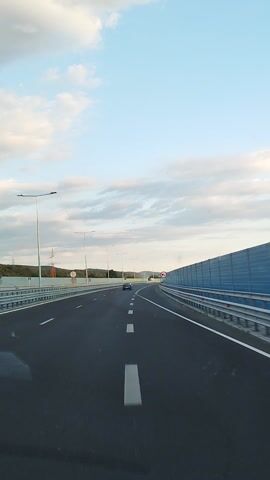 This media file is a first-person perspective video of a car driving on a highway. The road is paved with asphalt and marked with white lines. On the left side of the road, there are metal guardrails and lampposts. On the right side, there is a blue sound barrier and metal guardrails. In the distance, there are hills and trees. The sky is blue with scattered clouds, suggesting it is daytime, likely late afternoon or early evening.

The activity is driving on a highway. There are a few other vehicles visible on the road, including cars and a white van. There are no people visible in the media file, as the perspective is from inside the car.

Notable details include a red and white circular sign with a horizontal bar and a diagonal line through it, which typically indicates a prohibition. There is also a green highway sign with white text and numbers, possibly indicating a route number or exit.

Visible text:
- A green highway sign with white text that appears to include "A1".
- A red and white circular sign indicating a prohibition.