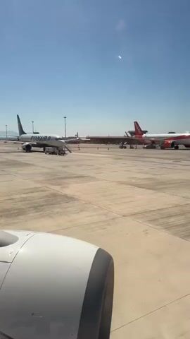 The video shows the tarmac at an airport on a sunny day. A white Ryanair jet is parked to the left, and a white and orange EasyJet jet is parked to the right, closer to the camera. Airport personnel and vehicles are working near the EasyJet plane. The video is taken from the window of another plane, with the engine visible in the foreground.