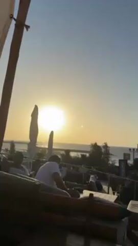 The video shows a beachside scene at sunset. In the foreground, there are wooden lounge chairs with cushions, partially under a fabric awning supported by wooden poles. Two men are sitting on the chairs, silhouetted against the setting sun. Beyond them, there's a view of the ocean, a beach, and some vegetation. White closed umbrellas are interspersed between the people and the water. A sign with the word "ACAI" is visible. The sun is a bright circle in the sky, partially obscured by the umbrellas. The sky is a light blue gradient, fading to a hazy orange near the horizon. People are scattered throughout the scene in various states of relaxation.