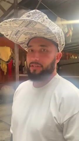 A man with a beard and a patterned cap is looking at the camera. He is wearing a white t-shirt. The background appears to be an indoor market or workshop with some goods hanging and displayed. The lighting is somewhat dim and artificial. The man smiles briefly.