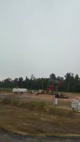 The video displays an oil pump jack operating in an open, rural landscape under an overcast sky. The scene is likely located in Ровенскаслабодскі сельскі Савет, Belarus, based on the provided context. The oil pump jack, a prominent red and metallic structure, is actively pumping, indicated by its rhythmic up-and-down motion. To the left of the pump jack, a large white horizontal tank is visible, likely used for storage in the oil extraction process. The surrounding area is a mix of cleared land and a treeline in the background, suggesting an industrial site within a natural setting. The weather appears to be cloudy, contributing to a diffused, neutral lighting. The overall impression is one of industrial activity in a relatively remote, natural environment. There is no visible text in the frame. The time of day is not precisely determinable due to the overcast conditions, but it appears to be daytime. No people are visible in the frame.