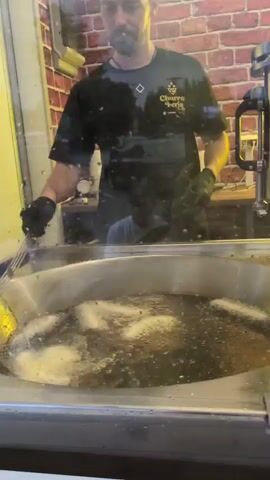 A person wearing black gloves and a dark t-shirt with a logo stands behind a large vat of bubbling oil. They are holding a metal mesh strainer over the oil, and several long, thin items are submerged in the oil, appearing to be frying. The background consists of a brick wall pattern and some equipment. The scene is viewed through a glass barrier.