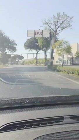 A POV shot from inside a vehicle shows a road on a sunny day. A signpost with "LAS" and other text is visible on the right side of the road. Trees line the road, and a large overpass or bridge structure can be seen in the distance. The vehicle appears to be driving on a paved road with lane markings. A speed limit sign of 50 is visible on a pole.