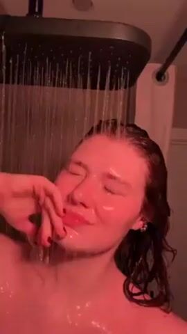 A fair-skinned young woman is shown from the chest up, standing directly under a square, dark-colored shower head. She has her eyes closed and is rubbing her scalp and face with both hands. The water is cascading directly onto her face and body. The lighting is red, which casts a pinkish hue over her skin and the water. A shower curtain can be seen behind her. The camera angle is looking upwards towards her face.