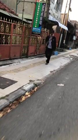 A person walks down a sidewalk in Kludan, Indonesia. The individual is wearing a dark jacket over a plaid shirt and dark pants. They are also wearing a mask, and carrying a dark bag over their shoulder. The person is walking away from the camera, and their face is not clearly visible.

The sidewalk is made of concrete and has a patterned border. There are some fallen leaves on the ground near the curb, suggesting it might be autumn, though Indonesia's tropical climate does not experience traditional autumn seasons. The street is paved with asphalt and appears to be mostly empty.

Behind the sidewalk is a fence with decorative metalwork, and beyond that, a building with a large green banner hanging from it. The banner displays text in Indonesian and appears to be an advertisement for a business. The sunlight suggests it is either early morning or late afternoon, casting long shadows. The overall scene is calm and ordinary, depicting a typical street view in a residential or commercial area.

Visible text on the banner:
"TANGGULACIN"
"TRA" (partially visible)
"BIASAKAN HIDUP SEHAT JAUHI YANG JAHAT"
"DISETERIA KESEHATAN"
"081 335 878 134"
"DAPATKAN PROMO KHUSUS"