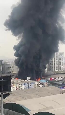 A massive plume of thick, black smoke billows into the sky from a fire engulfing a building. The fire is visible at the base of the smoke column, with orange flames licking upwards. Several multi-story buildings surround the burning structure, with one tall, white building prominently visible to the right. In the foreground, a portion of a lighter-colored roof or structure is visible, along with the top of a dark, rectangular object, possibly a billboard or a structural support. The sky is overcast, and the overall scene depicts an urban environment impacted by a significant fire.