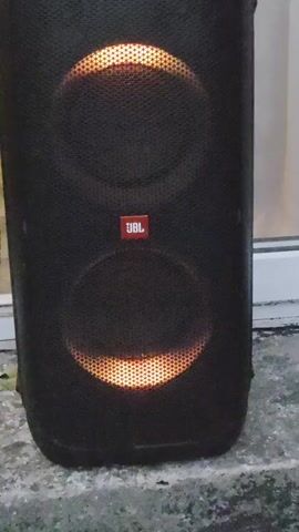A tall black JBL speaker with two circular lights in the center is shown. The lights are orange and yellow and appear to be pulsing. The speaker is sitting on a gray surface, possibly concrete, with some blurry objects in the background. The camera angle is straight on and the lighting is dim, with the speaker's lights being the brightest elements in the frame.