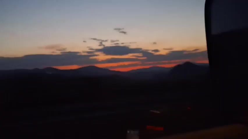 The video begins with a wide shot of a dusky sky with orange and red hues bleeding across the horizon, suggesting either sunrise or sunset. Silhouetted mountains stretch across the landscape, their peaks softened by the fading light. Scattered clouds drift above, catching the last rays of the sun. The foreground is dark, obscuring details but hinting at a roadway or open space. As the video progresses, the view shifts, zooming in slightly on the mountain range and the colorful sky. A few faint lights, possibly from distant vehicles or structures, are visible in the lower right portion of the frame. The overall impression is of a serene, twilight landscape.