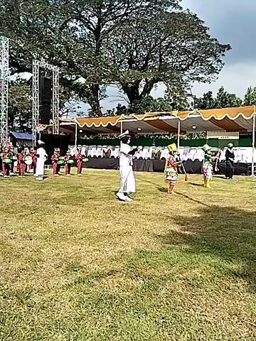 On a grassy field, a marching band in red and green uniforms performs in front of a stage. The stage has a white frame and a large banner with writing in front of it. Two tall metal towers stand on either side of the stage. Behind the stage is a large green tree and several white tents with orange awnings. Several people in white sit at the tents. In front of the marching band are two individuals in white outfits, directing the band. There is also another person in a costume with green, orange and red in front of the band.