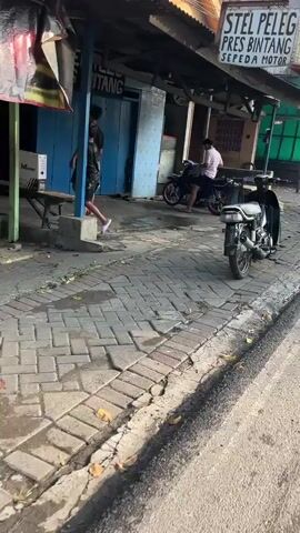 The video shows a street scene with several people and motorcycles. In the foreground, a motorcycle is parked on the right side of the frame. The pavement is made of interlocking bricks. In the middle ground, two young men are standing near a blue-doored building with a sign that reads "STEEL PRES BINTANG". One man is wearing a black t-shirt and shorts, and the other is wearing a black t-shirt and patterned shorts. To the right, a person in a pink shirt is sitting on a motorcycle. The background shows more of the building and a cluttered outdoor area. The lighting suggests it is daytime, possibly late afternoon.