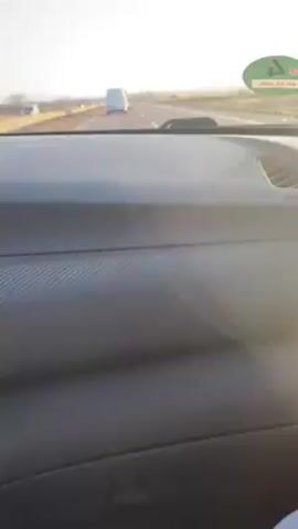 The video shows a view from the dashboard of a car driving on a highway. A white van is visible in the distance, driving in the same direction. Other cars are also visible on the road. The sky is blue and clear. The landscape beside the highway is mostly flat with some vegetation. The dashboard is dark grey and has a textured pattern. A round green sticker is visible on the windshield.
