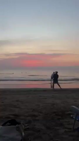 The video shows a beach scene at sunset. The sky is a mix of pink, orange, and grey colors. In the foreground, there is dark, sandy beach. A purse is lying on the ground and the corner of a blue chair can be seen. Further towards the ocean, two people are visible. One person stands in place, while the other is walking into the water. There are a few other figures in the distance, swimming in the ocean. The waves are small and gently breaking on the shore, reflecting the colors of the sunset. The overall scene appears calm and peaceful.