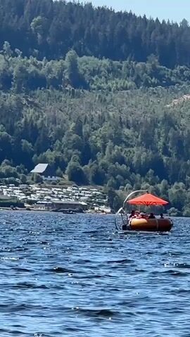 A group of people are on a circular, orange, inflatable boat with an orange umbrella providing shade. The boat is on a large body of water, likely a lake, with waves visible. In the background, there is a densely wooded hillside with a few buildings at the base. The sky is clear and blue. The boat appears to be moving slowly through the water.