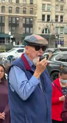 An older man with a long white beard, sunglasses, a blue collared shirt, a navy blue vest with red trim, and a multi-colored flat cap is speaking into a small black handheld device, likely a microphone or speaker. He is seen from the chest up, positioned slightly to the left of the frame's center, with his mouth open as if mid-sentence. To his right, partially visible, is a woman wearing a red jacket and a face mask, looking down. Another individual in a dark red top is partially visible to the man's left. The background features several multi-story buildings, characteristic of an urban environment, and parked cars, suggesting a city street or public square. The lighting appears to be overcast.
