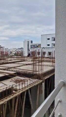 The video captures a construction site from a high vantage point. In the foreground, the exposed rebar and wooden scaffolding of a building under construction are visible. Several workers are present on the structure, with two individuals appearing to be engaged in work, and one standing and talking on a mobile phone. In the background, partially completed buildings and apartment complexes are seen under a cloudy sky. A person in a pink garment is seated on a balcony of a building in the distance.