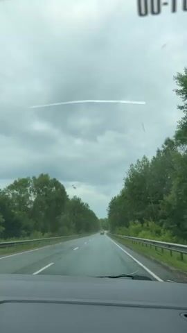 A view from inside a car driving on a wet asphalt road, flanked by trees on either side. The sky is overcast with a faint contrail visible. Several cars are ahead on the road.