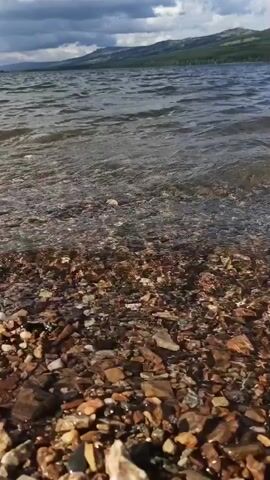 A body of water is viewed from a shoreline. The water has small waves and is rippling. The shoreline is made of small, multicolored rocks. In the background, there are hills and mountains covered with green trees. The sky is cloudy and overcast.