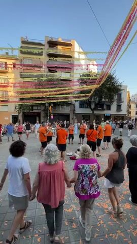 In a town square, a brass band in orange uniforms plays music as people gather around. Some watch from balconies of the surrounding buildings adorned with colorful streamers. In the foreground, four women hold hands in a circle, facing the band. The ground is covered in confetti. The scene appears to be a festive outdoor event.