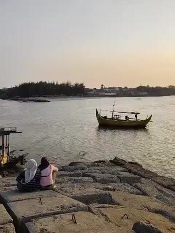 This is a video of two people wearing hijabs, sitting on concrete blocks overlooking a body of water. A yellow boat is in the water to the right. There is land with trees and buildings in the distance. The sky is clear and light.