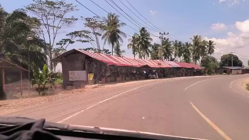 The video shows a view from inside a car driving along a road with a row of market stalls on the side. The stalls are covered with red and white striped awnings, and various goods are displayed in front of them. There are a few people visible in and around the stalls. Palm trees and other greenery are visible in the background. The sky is blue with some clouds. Power lines run along the side of the road.