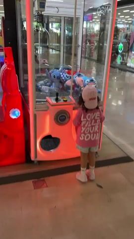 A young girl wearing a pink t-shirt and a pink baseball cap stands in front of a claw machine filled with Stitch plush toys. She is looking at the machine, and then turns her head to look at the camera. The claw machine is illuminated with pink lights. The background appears to be a shopping mall.