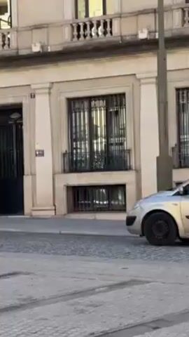 The video shows a panning shot of a European city square. It starts with a car parked in front of a light beige building, then pans to a larger, ornate building with flags, and a worker sweeping in front. The camera then dips, revealing the back of a cell phone with a beaded charm, before panning up to a row of tall buildings in red, yellow, and beige, with advertisements on their roofs. The sky is clear and blue.