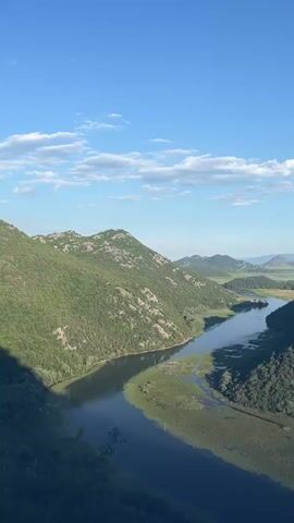This is a vertical video clip filmed from a high vantage point looking down on a winding river. The river flows through a valley with steep, tree-covered mountains on either side. The water is dark and reflective, with green vegetation growing along its banks. The sky is a clear blue with a few scattered white clouds. The sun is shining brightly, casting shadows on the landscape. The overall scene depicts a beautiful and serene natural landscape.