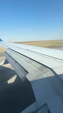 The video shows a view from an airplane window, focusing on the wing and the runway below. The wing is primarily grey with some visible panels and rivets. The flaps on the trailing edge of the wing are extended downwards, indicating the aircraft is either on the ground, preparing for takeoff, or has just landed. The runway is visible in the distance, appearing as a paved strip with markings. The surrounding environment is a flat, open landscape with sparse vegetation and a clear blue sky. There are no people or other objects clearly visible in the frame. The camera appears to be stationary, likely mounted inside the aircraft.