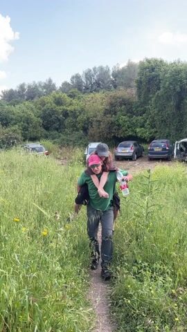 A man is giving a woman a piggyback ride. The man is wearing a green T-shirt, dark pants, a pink baseball cap, and sunglasses. He is holding a bottle of water. The woman is wearing a brown shirt and a black baseball cap. They are walking along a dirt path through a field of tall grass. Behind them, there is a line of cars parked in a clearing. In the background, there is a lush forest and a partly cloudy blue sky.