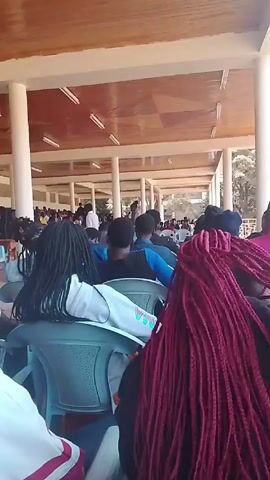 The video shows a large gathering of people, mostly Black, sitting in rows of chairs under a covered open-air structure. The structure has a wooden ceiling supported by white pillars. People are facing a stage area at the front, where some individuals are standing or moving around. Some people are wearing light-colored clothing, others are wearing darker colors. Several people in the foreground have braids of different colors. The overall setting looks like an outdoor event or assembly.