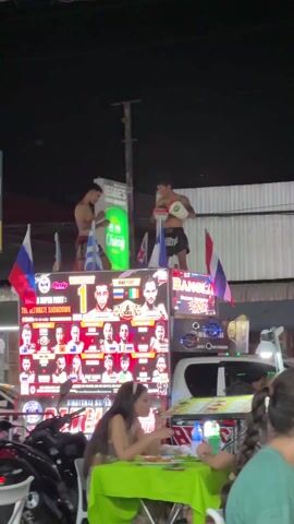 The video depicts a nighttime street scene in what appears to be a tourist area in Thailand. In the foreground, people are seated at tables, possibly dining. A large, illuminated sign is mounted on a vehicle, advertising an MMA fight night scheduled for Wednesday. Above the sign, two shirtless men in shorts are engaged in a sparring match or performance. The sign is decorated with flags from various nations, including Russia, Greece, and Thailand. The overall atmosphere suggests a lively and bustling entertainment district.