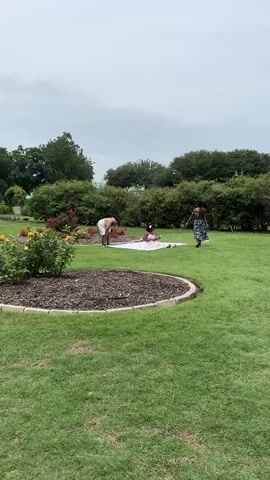 The video shows three women in a park on a cloudy day. One woman is sitting on a white blanket spread on the grass. She is wearing a pink dress with a floral pattern. She waves at the camera. Another woman is standing next to the blanket, wearing a white dress. The third woman is standing a little further away, wearing a blue dress with a floral pattern. The area is landscaped, with well-maintained grass and flower beds. There are trees and bushes in the background. Other people can be seen walking in the distance near a fence with arches over it.