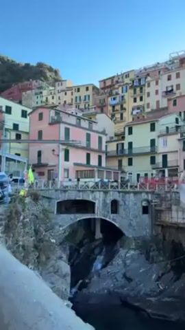 The video shows a picturesque Italian coastal village, likely Manarola in Cinque Terre, built on a cliffside. Colorful buildings in pastel shades of pink, yellow, and green are stacked upon each other, rising from a rocky coastline. A stone bridge with an arched opening spans over a narrow ravine through which water flows. A portion of the coastline is rugged and rocky, with construction or maintenance appearing to be in progress near the bridge. The sky is clear and blue, with a visible contrail from a passing aircraft. A few people can be seen in the video, either walking or standing by the buildings.