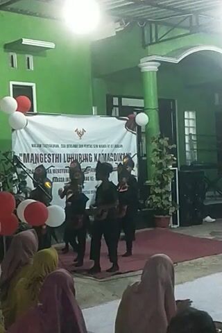 The video shows an outdoor event at night. A group of performers in dark costumes and masks are on a stage in front of a large banner. The banner has text in Indonesian and an image of the Indonesian coat of arms. There are balloons, including red and white ones, on the stage. A few people are seated in the foreground, watching the performance. The background appears to be a building with green walls and a white pillar. The lighting is artificial.