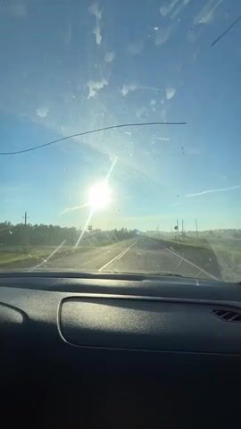 The video shows a view from inside a car, looking through the windshield. The sun is shining brightly in a clear blue sky, creating lens flares. A cracked windshield is visible, with some smudges and streaks. The car is driving on a paved road that appears to be in a rural or suburban area. Trees and utility poles are visible on either side of the road. The overall impression is one of a sunny day with a car in motion.