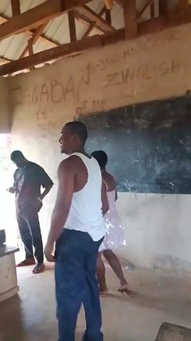 The video shows three individuals in what appears to be a classroom or an unfinished building. A man in a white tank top and dark pants is in the foreground, facing away from the camera and looking to his left, his right arm raised. To his right, a woman in a short, sparkly pink dress is dancing, facing away from the camera. She is holding a black object in her right hand. Behind the man in the white tank top, another person, likely male, is partially visible, also looking to the left. The environment features a dirt floor, a wooden ceiling, and walls with graffiti. A blackboard is mounted on the wall behind the woman. The lighting suggests it is daytime, with light coming from an unseen source to the left.