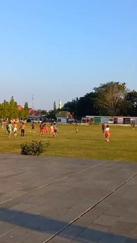 A group of young boys are playing soccer on a grassy field. They are wearing casual clothing, and some are shirtless. The game is informal, with no clear boundaries or referee. In the background, there are trees, buildings, and a clear blue sky. The sun is shining, casting long shadows across the field.